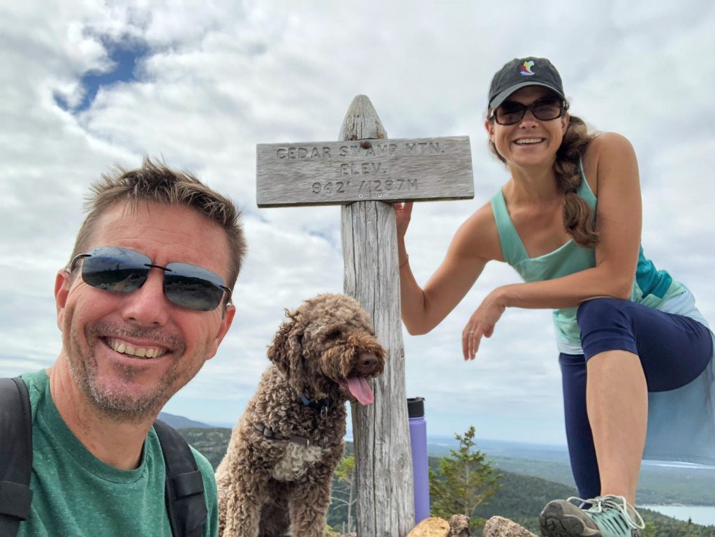Keith Tharp on left, Kate Harris on Right and their Dog Kalee in the middle, all posing near the summit sign for Cedar Swamp Mountain in Acadia National Park